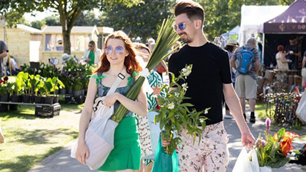 A woman and a man are walking past stall selling flowers. The woman has long red hair and is wearing a green dress and sunglasses. The man is wearing a black t-shirt and floral shorts and is wearing sunglasses. Both are carrying plants they have just purchased.