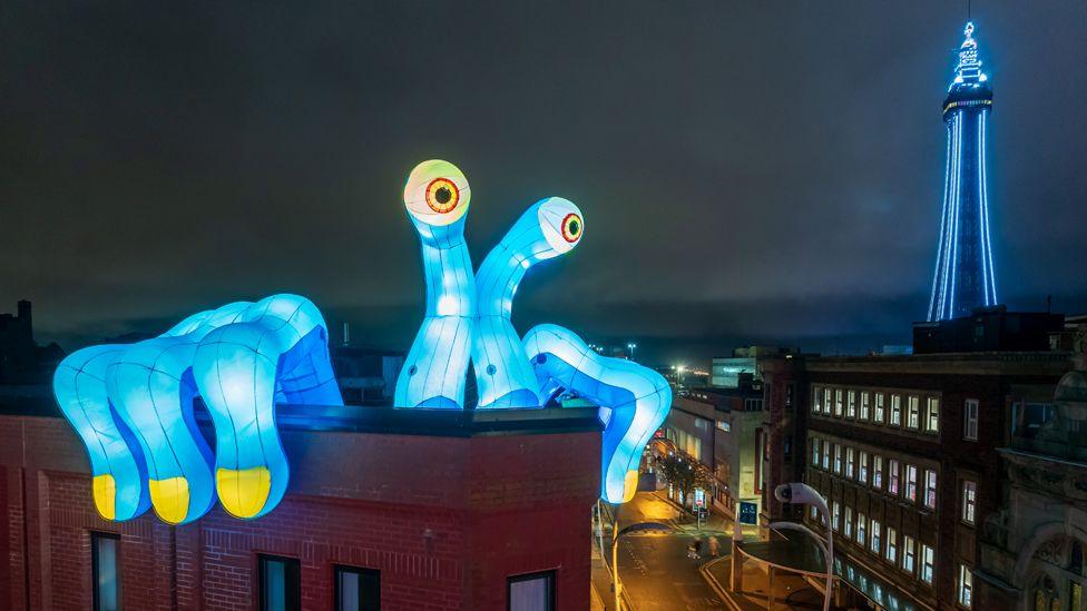 A blue inflatable monster with long fingers and eyes on stalks peers over the side of a building as Blackpool Tower shines against the night sky in the background