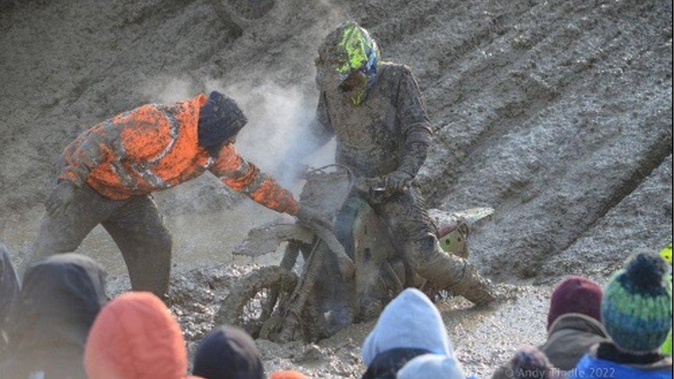 Rider and bike covered in mud are being helped through the mud by a marshall in orange hi-viz