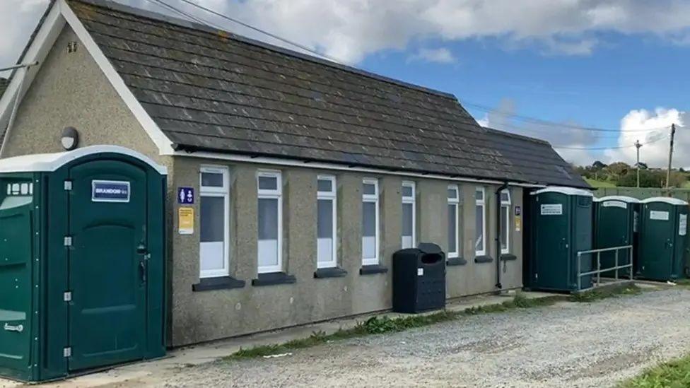 A pebble dashed single storey building with eight upright windows and a bin at the front along with a number of temporary loos each side of the building.
