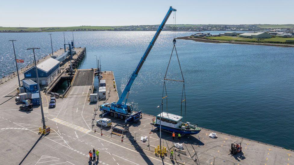 A small ferry being lifted by a crane from the deck towards a harbour with shimmering blue water