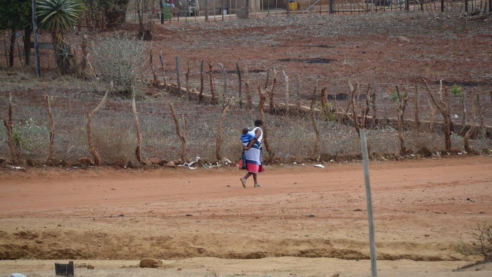 A woman walks through a village with a baby on her back