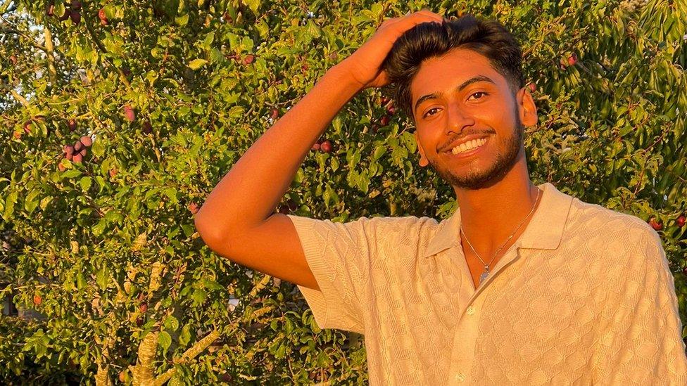 Kirthigan, a 19-year-old south Asian man, smiles at the camera with a hand in his short dark hair. Kirthigan, who has a short moustache and bears and brown eyes wears a short-sleeved white shirt and a silver necklace. He is pictured outside at sunset during "golden hour" in front of fruit trees.