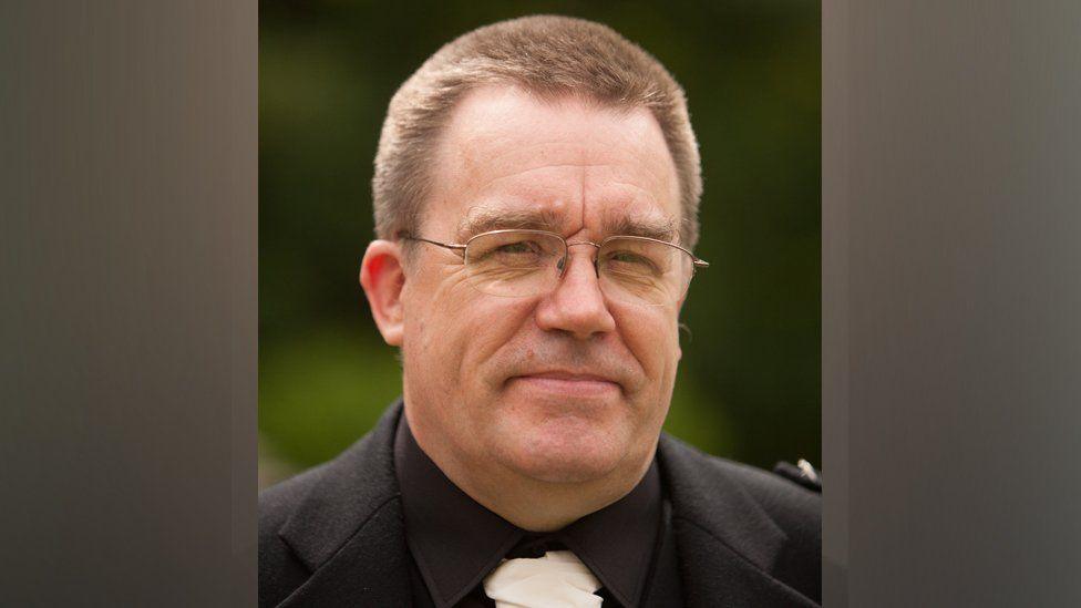 A man with short brown hair wearing a dark suit, white tie and glasses.