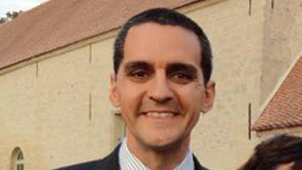 Richard Osman, who has short dark hair and is wearing a striped shirt and suit jacket and is smiling at the camera. A beige coloured building is behind him.