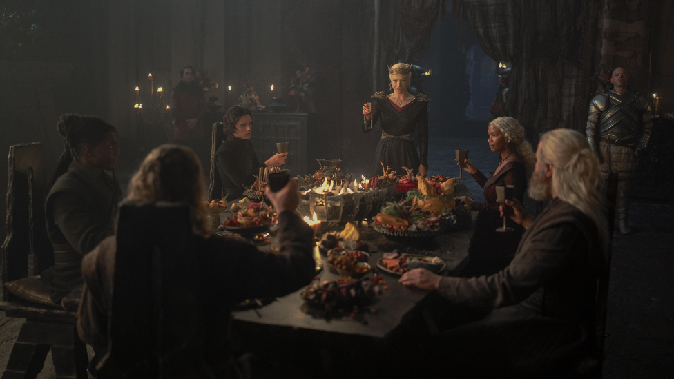 Group of people in House of the Dragon, sat around an elaborate feast raising their glasses to toast