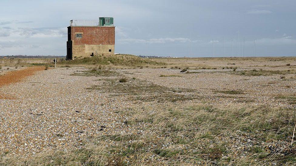 A Cold War era two storey brick built flat-roofed building standing against a blue-grey sky. It is surrounded by shingle. In the foreground on the right are some coastal grasses and other plants