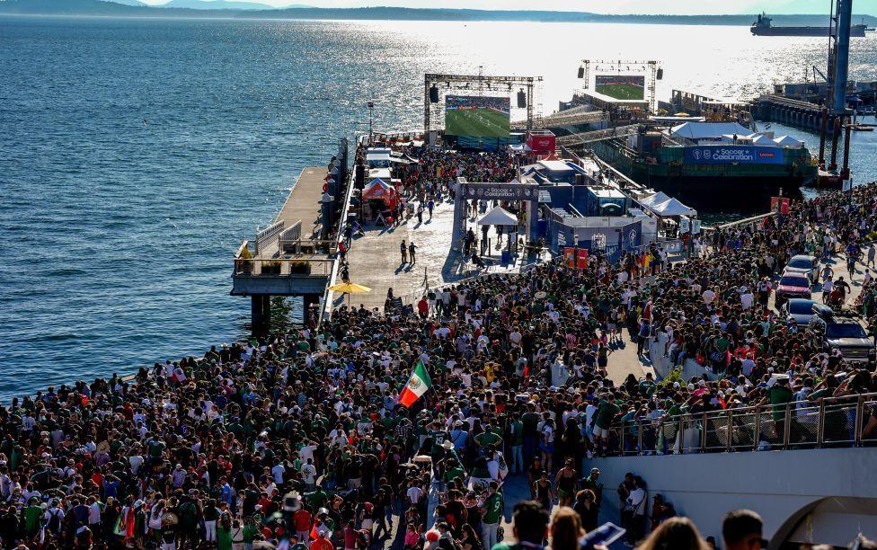 Crowds of people watching a big screen at the Seattle waterfront. The water in the harbour is glistening in the sunshine which is coming over the far horizon