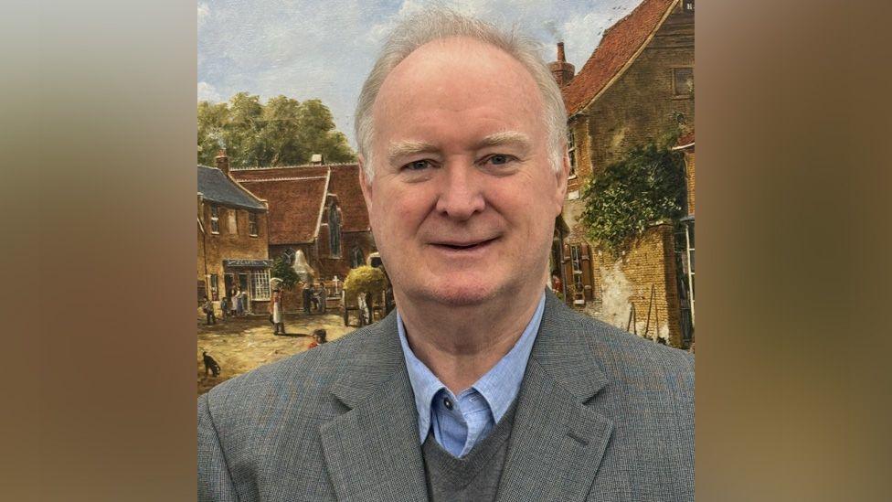 Russell Fowler in a grey suit and blue shirt standing in front of a painting