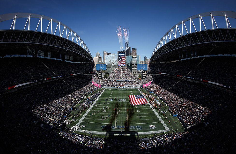 A panoramic view of Lumen Field during the NFL 2025 game between the Tampa Bay Buccaneers and Seattle Seahawks. The stadium is packed with fans, a large American flag is on the field, and fireworks rise above the scoreboard against a clear blue sky.