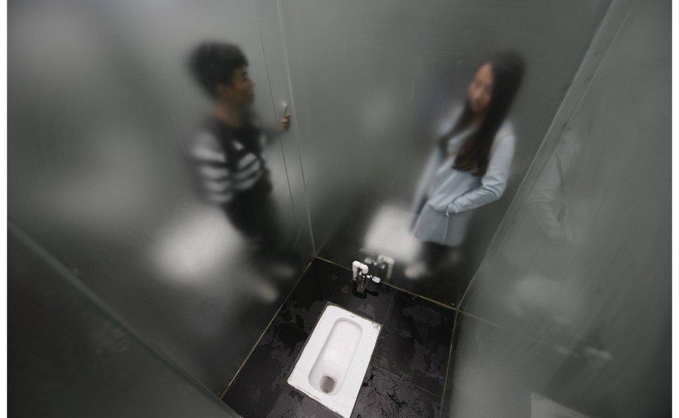 A squat toilet cubicle surrounded by lightly frosted glass, with a man on the other side of one glass wall and a woman on the other side of another glass wall. Changsha, China, 29 September 2016.