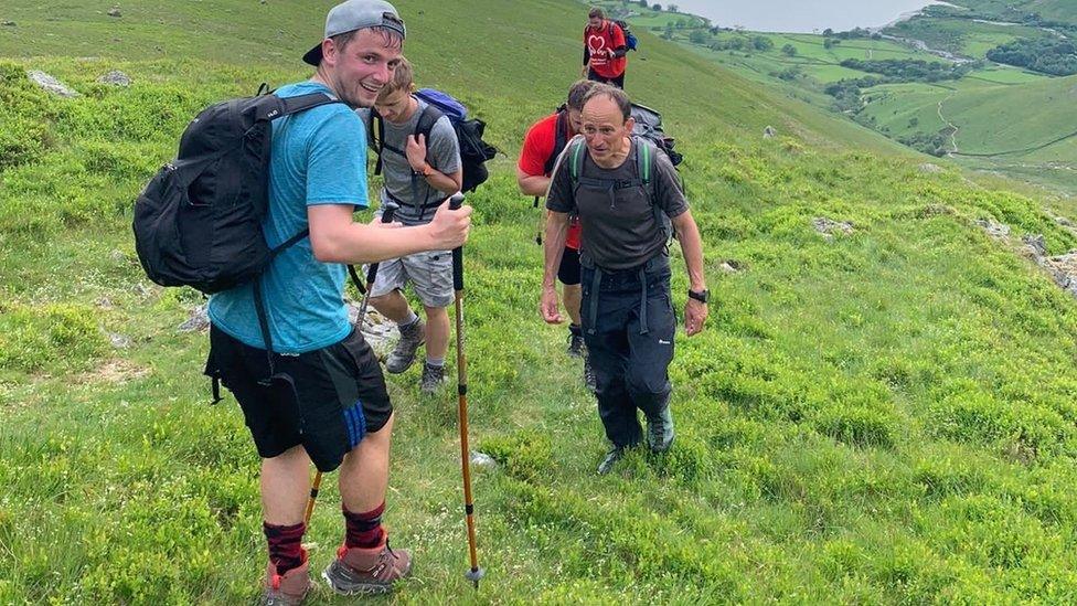 Ben Stewart walking up a hill backwards, looking over his shoulder and using a walking stick. Four men are walking up the hill behind him, facing towards the camera