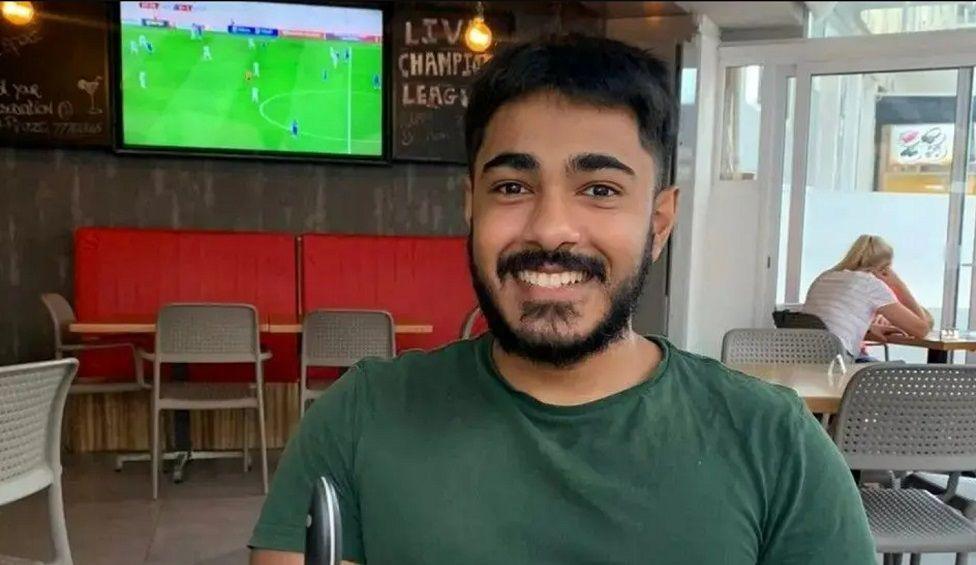 Anugrah Abraham wearing a green top and sat in a public eatery that has a television playing a football game