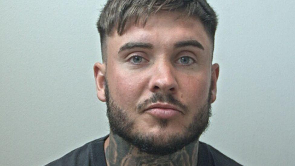 A police mugshot of Ryan Wellings, 30, who has short dark blonde hair and a short dark beard, and a tattoo covering most of the front of his neck. 