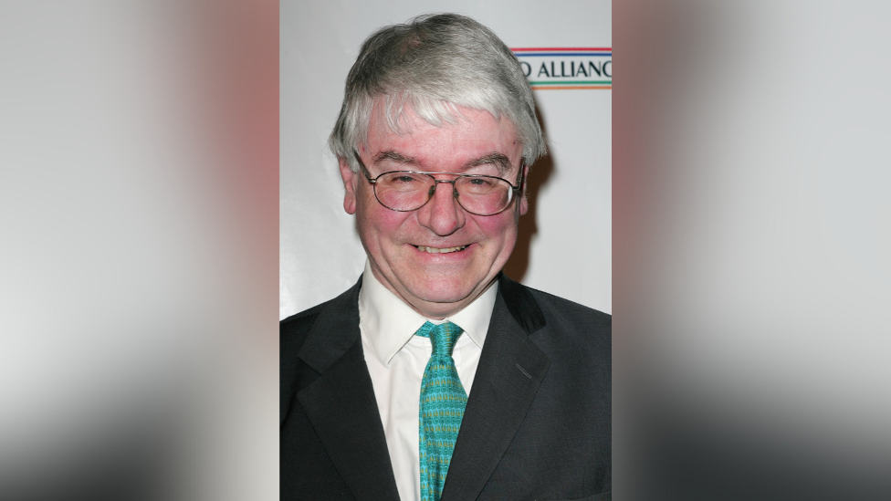 Mr Mansergh is smiling. He wore glasses and is wearing a suit with a green tie. 