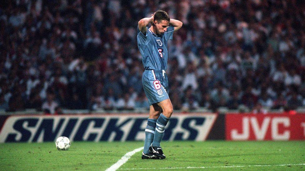 Gareth Southgate, a man in a blue football kit, stands on a pitch with his hands behind his head, looking defeated