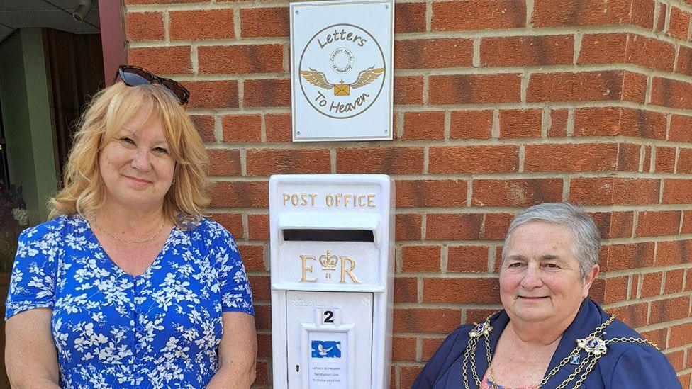 Sharon Sutcliffe has blond hair and sunglasses on top of her head. She is wearing a blue dress with a flower pattern and is standing on the left-hand side  of a white postbox, which is mounted on a brick wall with a sign above it. To the right stands Hull's Lord Mayor Councillor Cheryl Payne who has short grey hair and dark blue top with the silver mayoral chains around her neck.