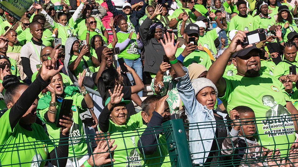 A crowd of people behind a fence, all wearing green T-shirts of the Patriotic Alliance, wave their hands in the air.
