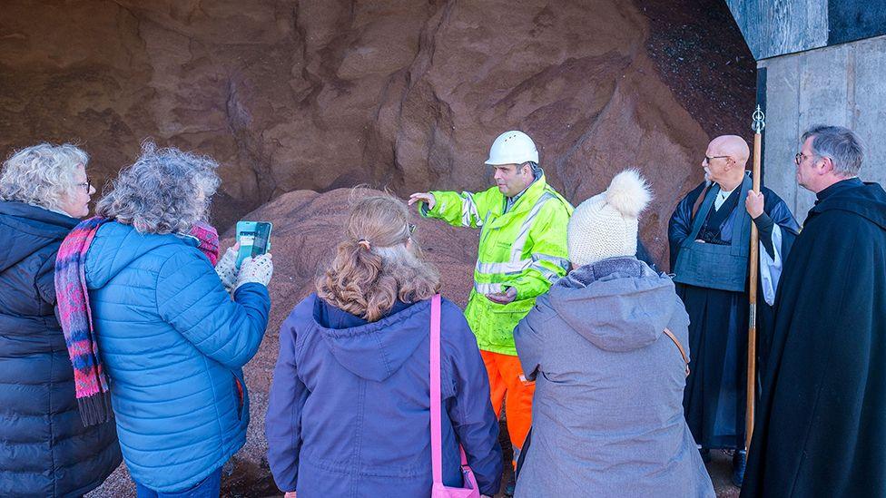 Members of the faith group with their backs to the camera having a tour of the gritting depot by a manager dressed in a white hard hat and high visibility clothing stood in front of a mound of brown salt