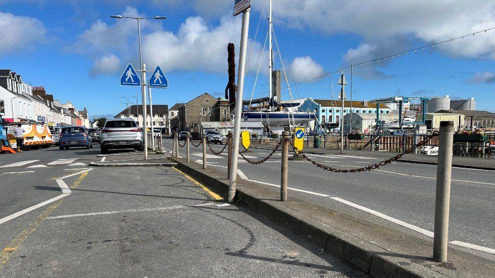 A photo which shows an area called The Bridge in Guernsey. There are metal poles which are acting as a fence in the middle of the photo, and two car parking spaces shown.