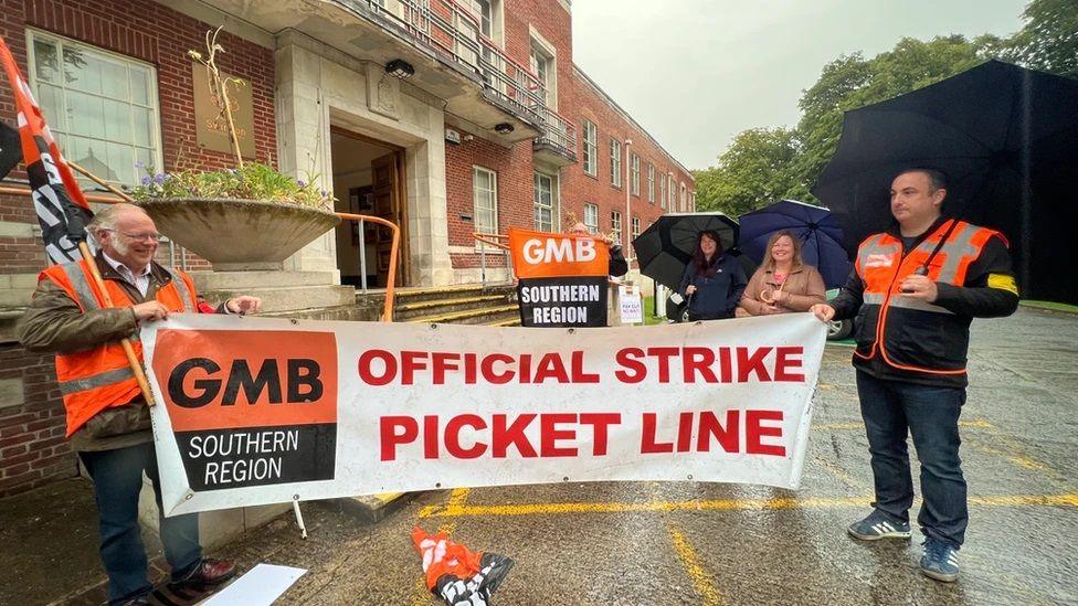 Two people holding a GMB Union strike picket line banner on a rainy day outside a large red brick building.