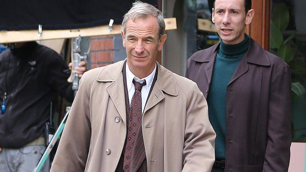 Robson Green during filming for the drama. He has short grey hair, and is wearing a beige overcoat over a pin-striped suit, white shirt and tie and appears to be walking towards the viewer. Behind him is a man wearing a dark jacket over a teal polo-neck jumper. Behind them can be seen a black screen held up by a man in a bomber jacket and jeans, whose head is shielded from the screen.