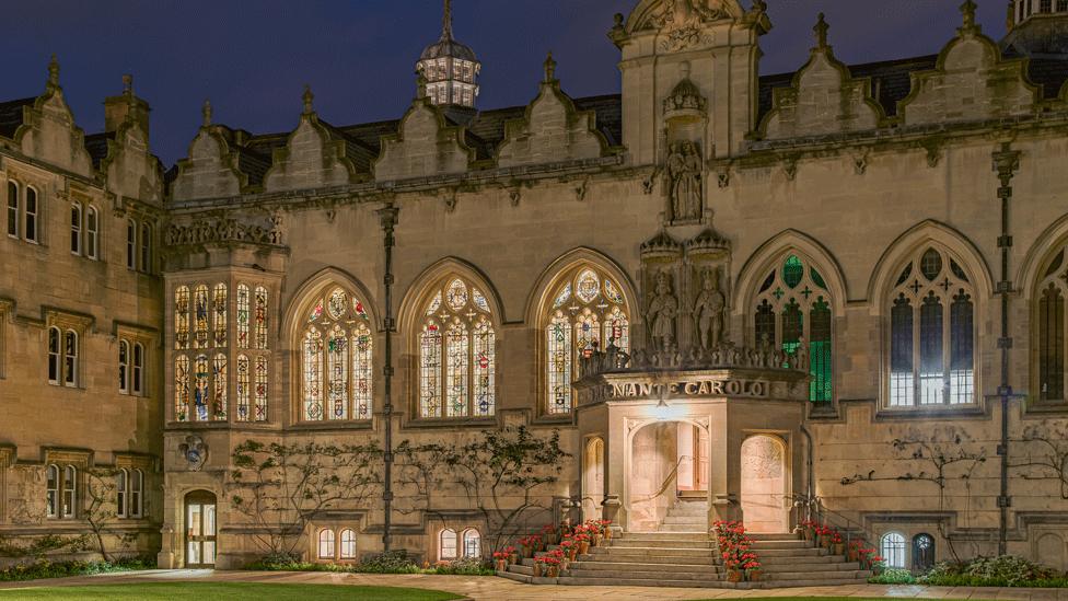 Oriel College First Quad at night time. The gothic building is lit up from inside, light glowing from stained glass windows. Steps leading up to a portico are lined with red geraniums.