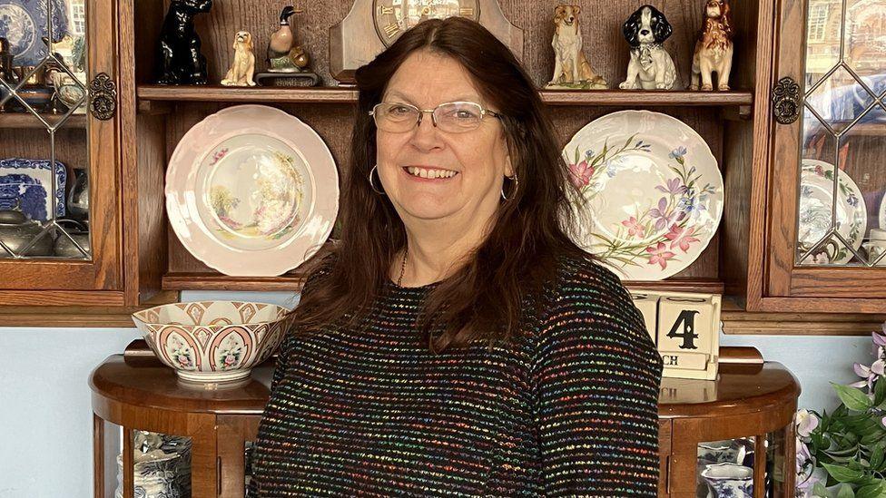 Sue Noades, with long brown hair, glasses and a multicoloured top in front of vintage ornaments