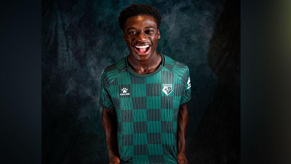 A young man wearing the new Watford away shirt and giving a huge smile, looking very energised. He is standing against a black background.