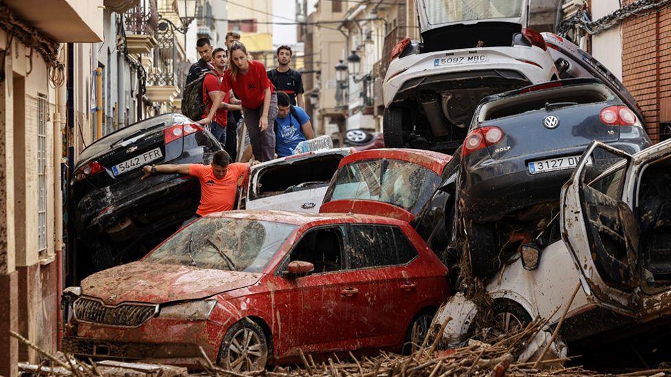 A group of people climb over piled up cars in a street in the flood-hit municipality of Paiporta, Valencia province