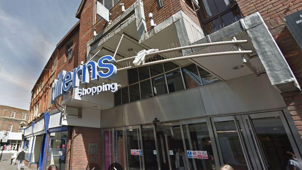 The sign Chilterns Shopping is seen from the right side in blue and white, over a set of glass and silver shopping centre doors at the main entrance. A brown/red brick building on a pedestrianised street, with heritage red brick row of shops to its left.