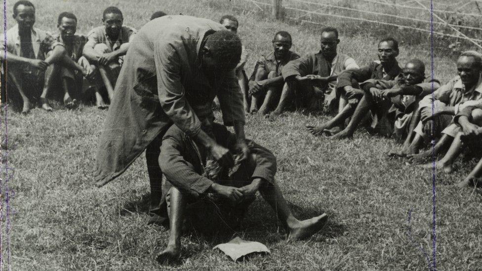 Government de-oathing ceremony, 1953 - A Mau Mau detainee sitting in the centre of a circle of men in a fenced compound is purified of the Mau Mau oath.