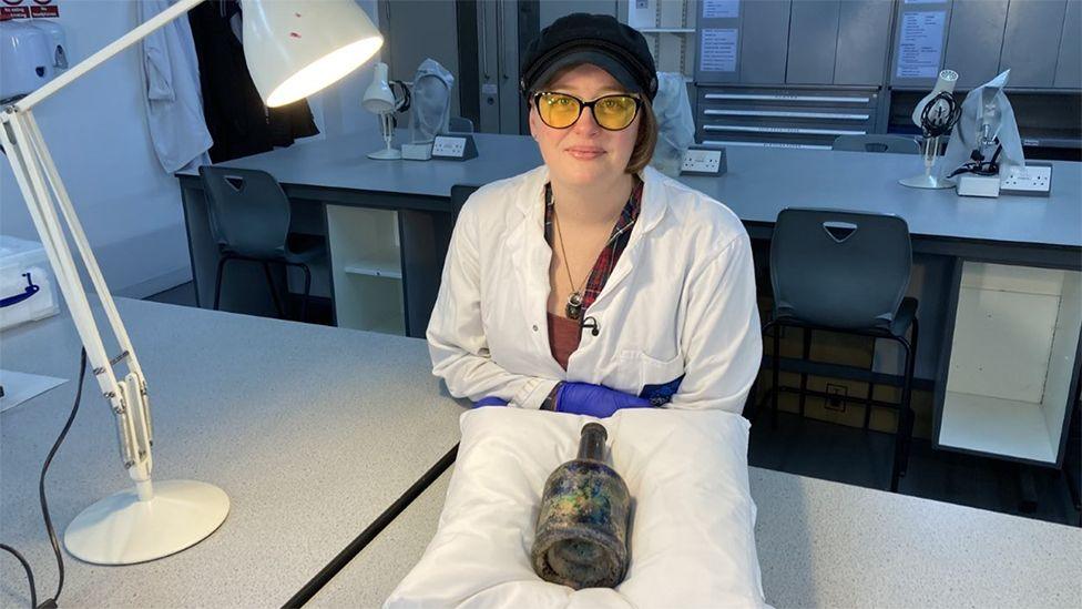 Zara Yeates sits at a laboratory table with the bottle on a cushion in front of her she is wearing a white lab coat, glasses and a blue cap