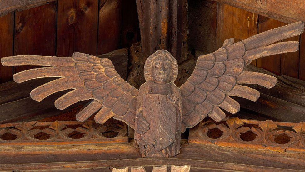 A wooden angel on the roof of St Wendreda's, March. Its wings our outstretched and it holding a shield in its hands. It is attached to a carved beam and above it can be seen the apex beam of the roof 