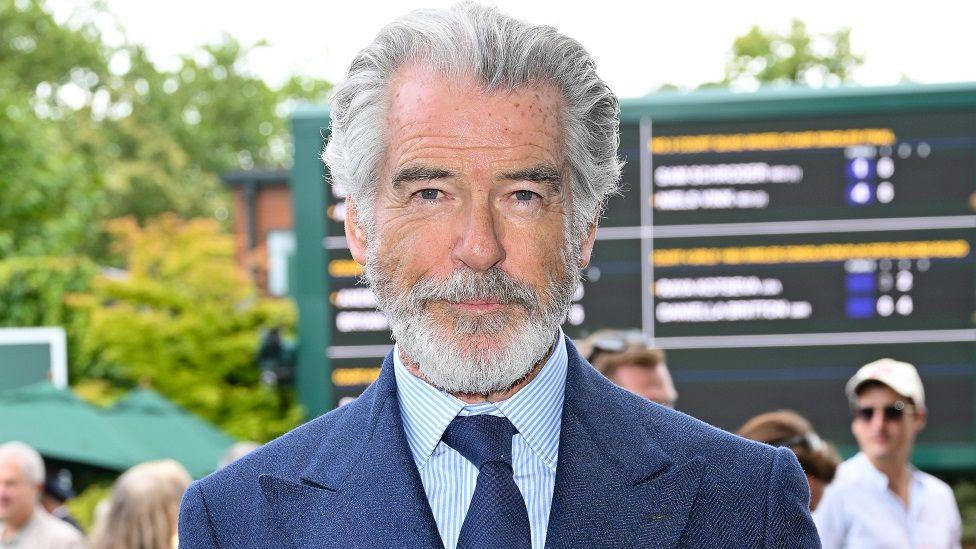 Brosnan posing for the camera at Wimbledon. He has grey hair and a grey beard