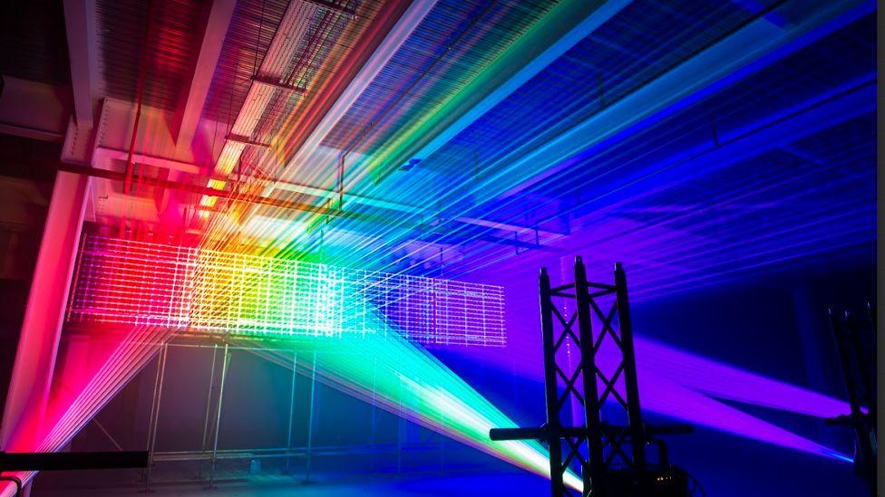 Two spotlights, one pink and one green, shine on to a dark wall, creating rainbow-coloured laser beams across the ceiling