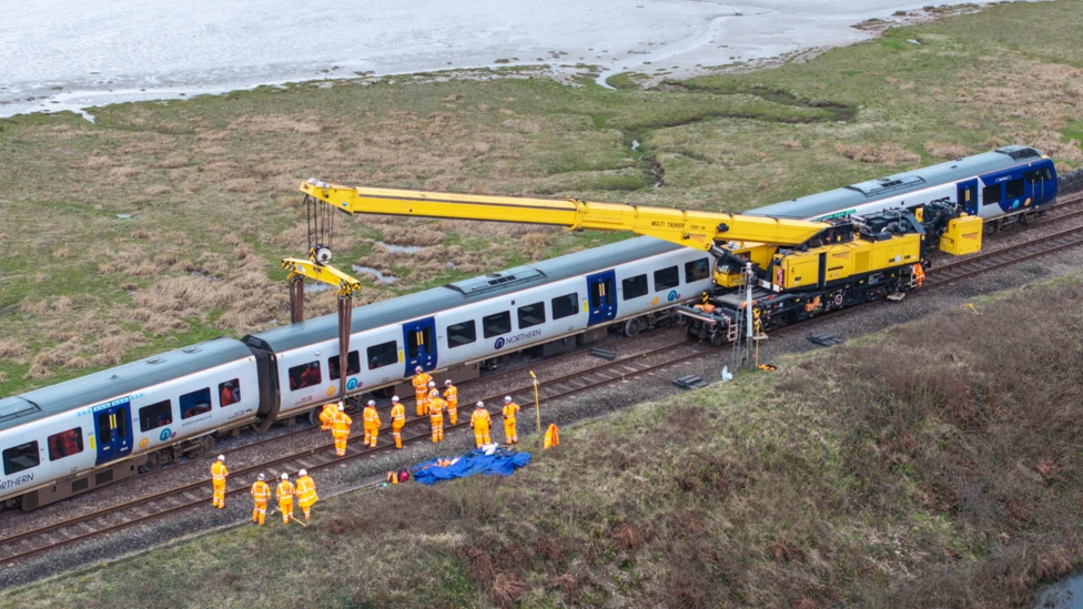 A specialist crane rerailing the Northern train at Grange-over-Sands. There are dozens of workers wearing bright orange uniform while the crane lifts the middle carriage. 