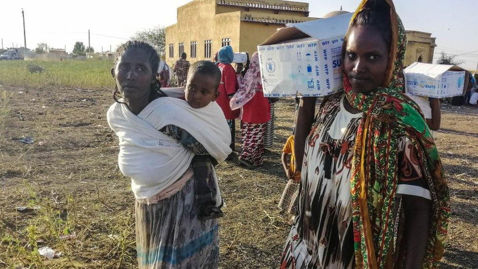 Two women, one holding a baby and the other with a box on her shoulder