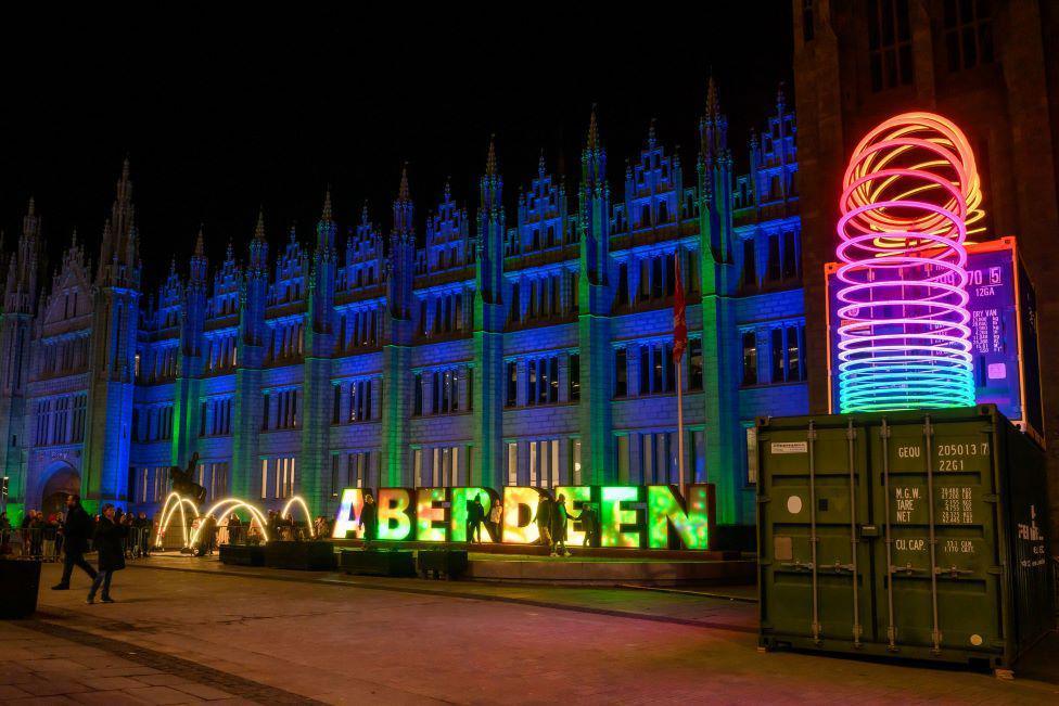 Impressive granite Marischal College building lit up blue, with the word 'Aberdeen' in block capitals also lit up, and a bright slinky-style object.