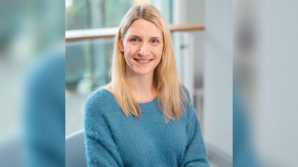 A portrait shot of a woman with shoulder length blonde hair and a fluffy blue jumper smiling at the camera. The background is blurred and it looks like she is sat in an office-type room.