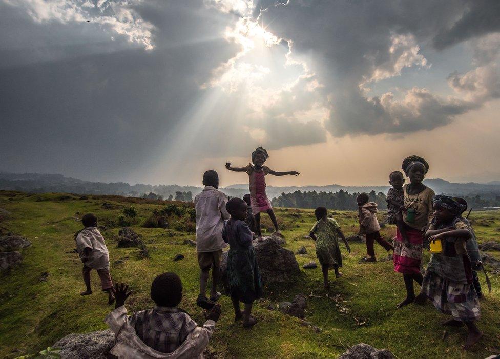 children in a green expanse, one jumping up and down on a rock, as the sun breaks through a cloud