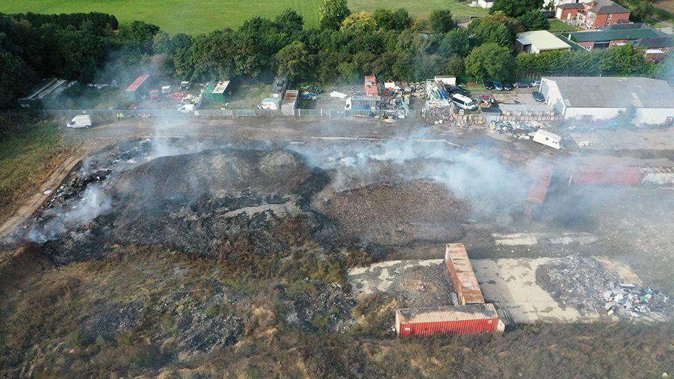 An aerial view of the site showing smoking piles of rubbish with shipping containers and other structures 