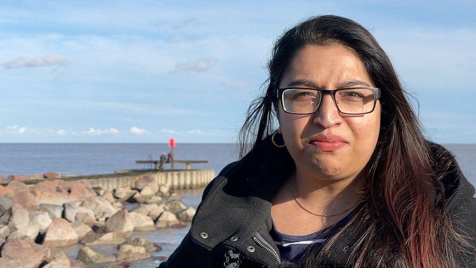 Nasima Begum stands on a seafront on a sunny day and smiles at the camera. She has long brown hair and wears glasses, hoop earrings and a black coat.