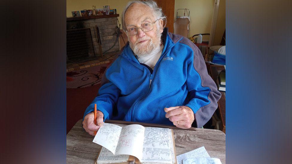 Wilfred sits at a desk in his living room with a book open in front of him that contains road maps. He holds an orange pen in one hand and looks at the camera. He wears a white t-shirt and blue and grey fleece, zipped up.
