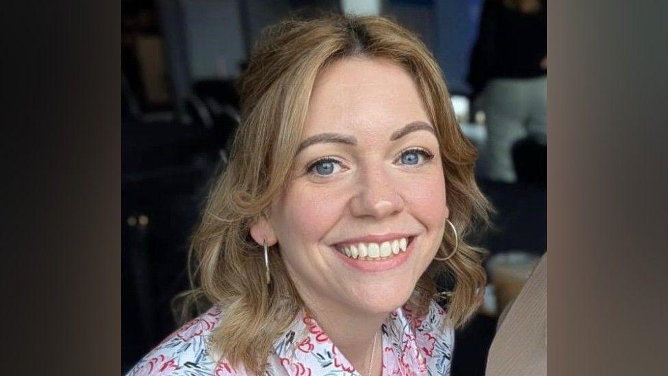 A woman with dark blonde hair is smiling at the camera, she is wearing a white top with pink and green flowers on it, and silver hoop earrings
