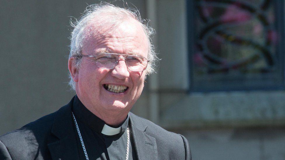 a smiling Bishop Donal mckeown in full clerical dress standing in front of a stained glass window