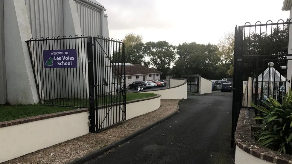A black school gate with a small sign which says Les Voies School on it. Behind it is a care park in front of a brown building.