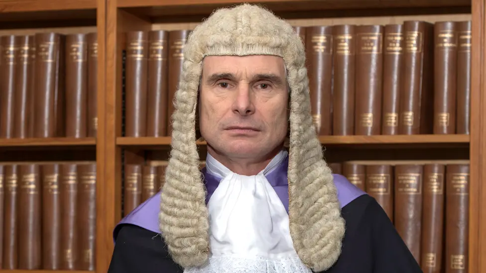 Judge Tolson in legal wig and gown in front of a brown bookcase