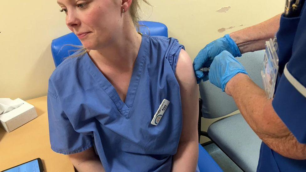 A woman dressed in blue medical scrubs being injected with the whooping cough vaccine by a nurse