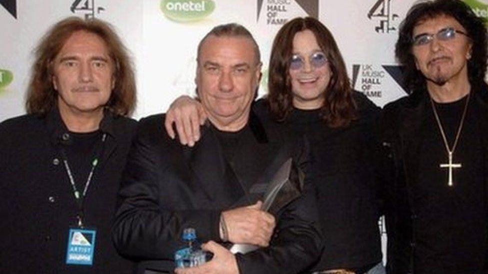 Black Sabbath band members smiling at the camera, all wearing black clothes.